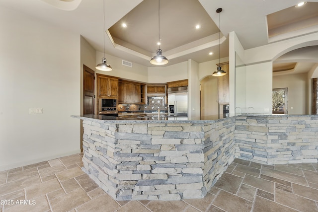 kitchen with a raised ceiling, built in appliances, dark stone counters, and pendant lighting