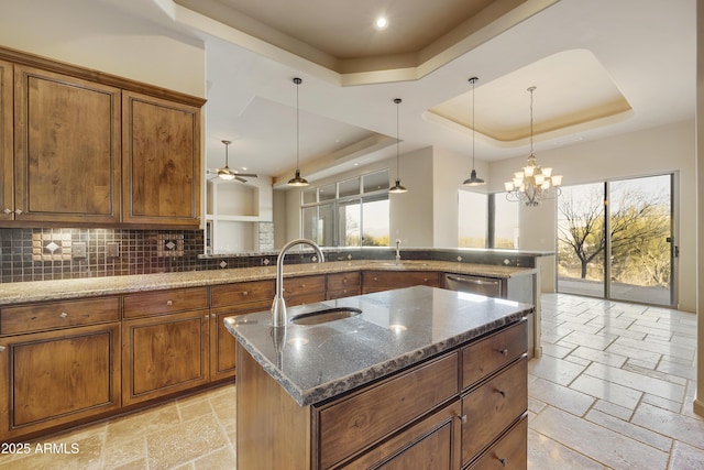 kitchen featuring dark stone counters, ceiling fan with notable chandelier, a tray ceiling, a kitchen island with sink, and hanging light fixtures