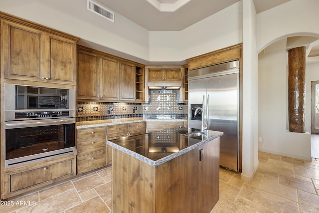 kitchen with stainless steel appliances, tasteful backsplash, a towering ceiling, dark stone counters, and a center island with sink