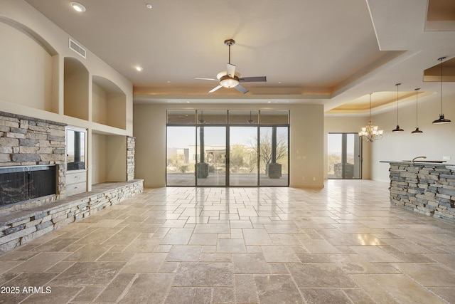 living room featuring a raised ceiling, a stone fireplace, plenty of natural light, and ceiling fan with notable chandelier