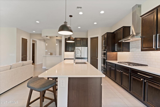 kitchen with a kitchen bar, wall chimney exhaust hood, stainless steel appliances, hanging light fixtures, and an island with sink