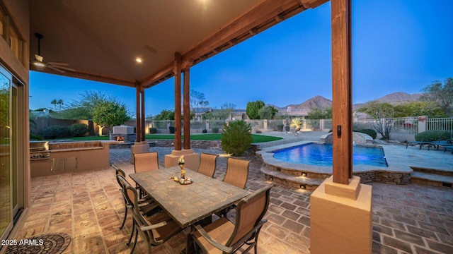 patio terrace at dusk featuring pool water feature, ceiling fan, area for grilling, a mountain view, and a fenced in pool