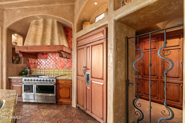 kitchen with light stone counters, premium range hood, range with two ovens, and backsplash