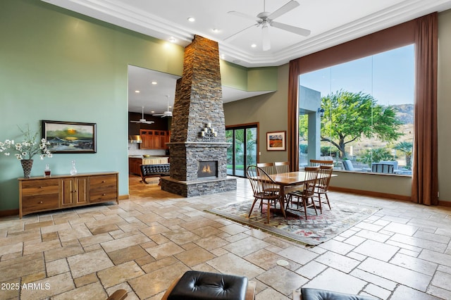 dining area with a towering ceiling, a stone fireplace, baseboards, stone tile flooring, and ceiling fan