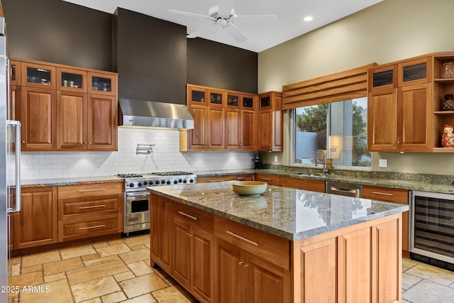 kitchen with stone tile flooring, wall chimney range hood, appliances with stainless steel finishes, a ceiling fan, and wine cooler