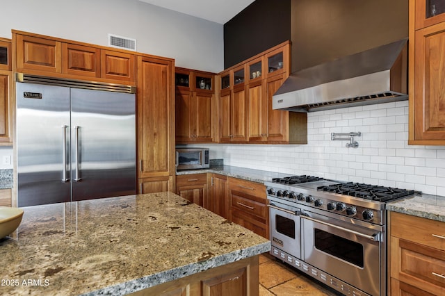 kitchen featuring light stone counters, high quality appliances, wall chimney range hood, backsplash, and visible vents