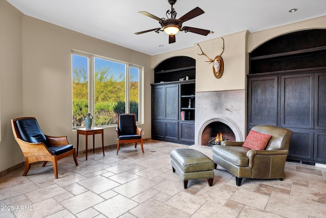 sitting room featuring built in features, ceiling fan, a premium fireplace, baseboards, and stone tile flooring