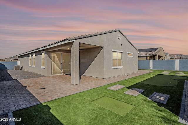 back of house at dusk featuring cooling unit, a fenced backyard, a lawn, stucco siding, and a patio area