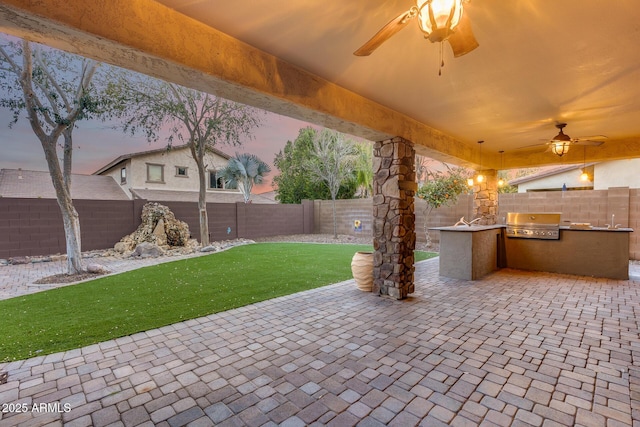 patio terrace at dusk featuring exterior kitchen, a grill, a yard, and ceiling fan