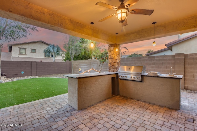 patio terrace at dusk featuring an outdoor kitchen, sink, a yard, ceiling fan, and a grill