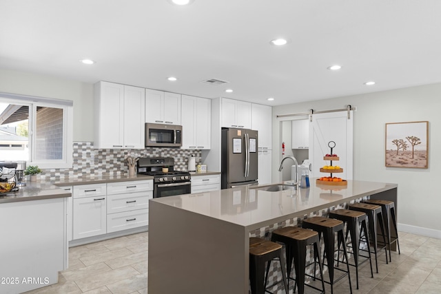 kitchen with sink, stainless steel appliances, an island with sink, white cabinets, and a barn door