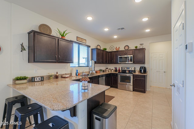 kitchen featuring stainless steel appliances, kitchen peninsula, a breakfast bar, light stone counters, and dark brown cabinetry