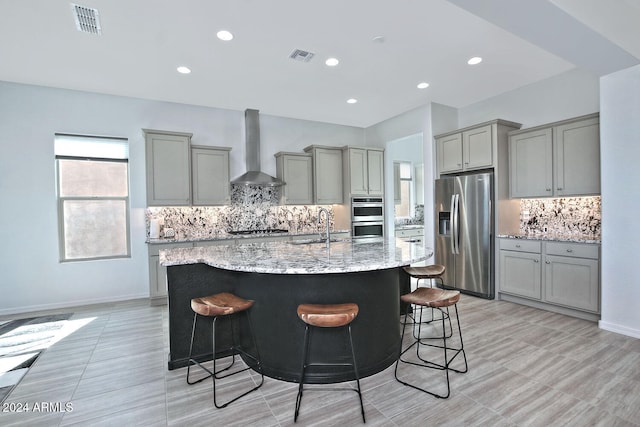 kitchen featuring wall chimney range hood, a center island with sink, a kitchen bar, gray cabinets, and stainless steel appliances