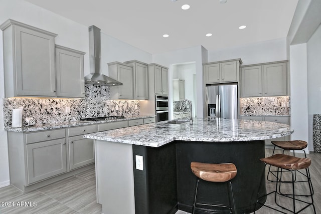 kitchen featuring wall chimney range hood, appliances with stainless steel finishes, gray cabinets, a breakfast bar area, and a kitchen island with sink