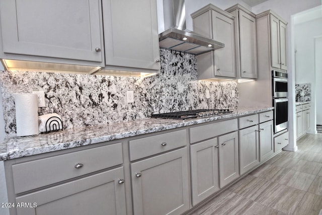 kitchen with wall chimney range hood, backsplash, light stone countertops, gray cabinets, and appliances with stainless steel finishes