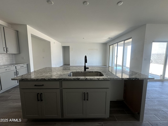 kitchen with dark stone countertops, tasteful backsplash, a sink, gray cabinetry, and an island with sink