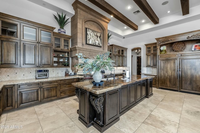 kitchen featuring a kitchen island with sink, decorative backsplash, light stone countertops, beamed ceiling, and dark brown cabinetry