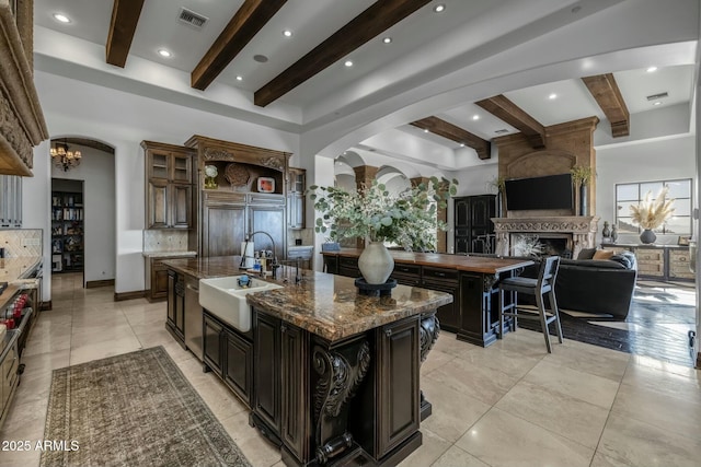 kitchen with dark brown cabinets, a large fireplace, sink, a large island with sink, and beamed ceiling