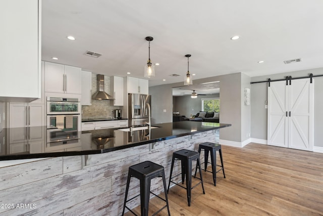kitchen with dark countertops, a barn door, wall chimney exhaust hood, double wall oven, and visible vents