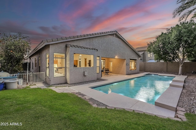 back of house at dusk with a fenced backyard, a lawn, a fenced in pool, stucco siding, and a patio area