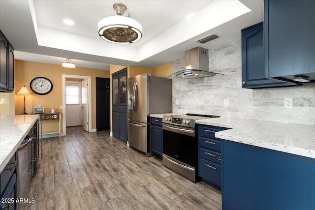 kitchen featuring stainless steel appliances, a tray ceiling, blue cabinetry, and wall chimney exhaust hood