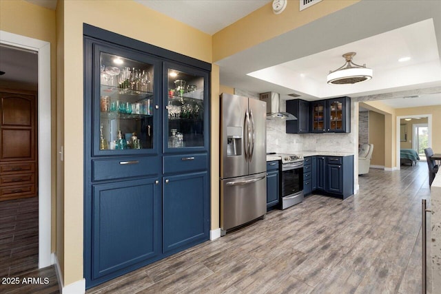 kitchen featuring wall chimney range hood, blue cabinetry, backsplash, hardwood / wood-style floors, and stainless steel appliances