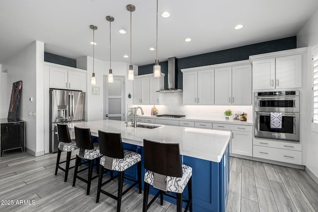 kitchen featuring a sink, wall chimney exhaust hood, appliances with stainless steel finishes, an island with sink, and white cabinets
