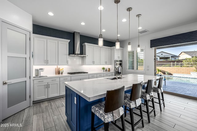 kitchen with wall chimney range hood, light wood-style flooring, a kitchen island with sink, a sink, and recessed lighting