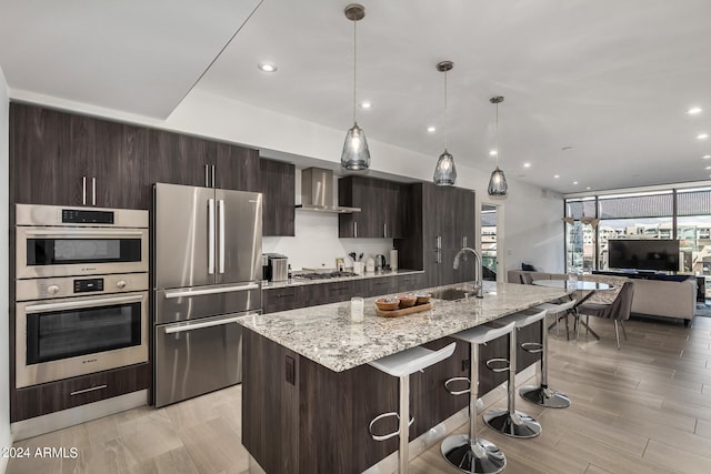 kitchen featuring a kitchen island with sink, appliances with stainless steel finishes, a kitchen breakfast bar, and wall chimney range hood