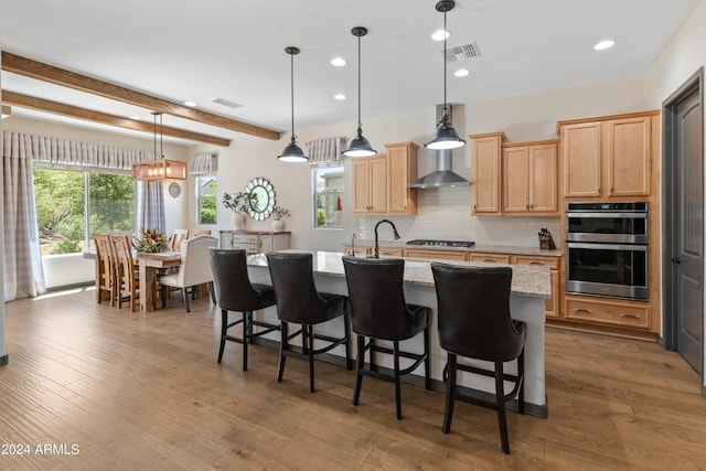 kitchen featuring pendant lighting, stainless steel double oven, an island with sink, and wall chimney range hood