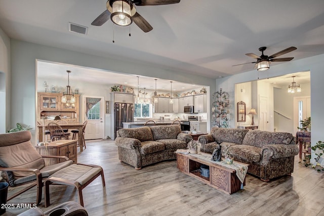 living room featuring light wood-style flooring, ceiling fan with notable chandelier, and visible vents