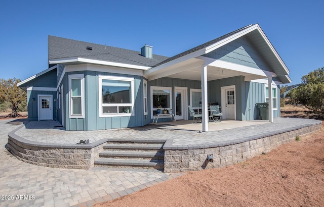 rear view of house with board and batten siding, a shingled roof, and a patio