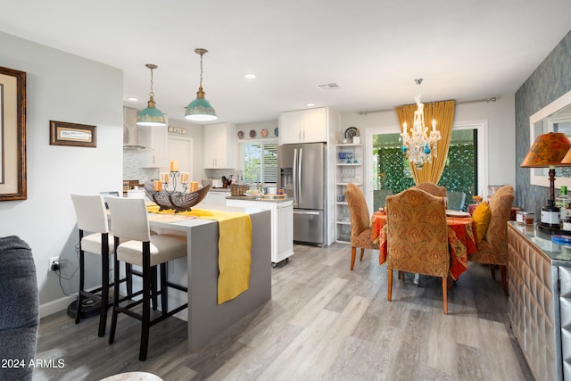 kitchen with white cabinets, hanging light fixtures, wall chimney exhaust hood, stainless steel fridge, and light wood-type flooring