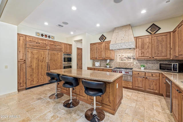 kitchen featuring appliances with stainless steel finishes, backsplash, light stone counters, custom range hood, and a center island