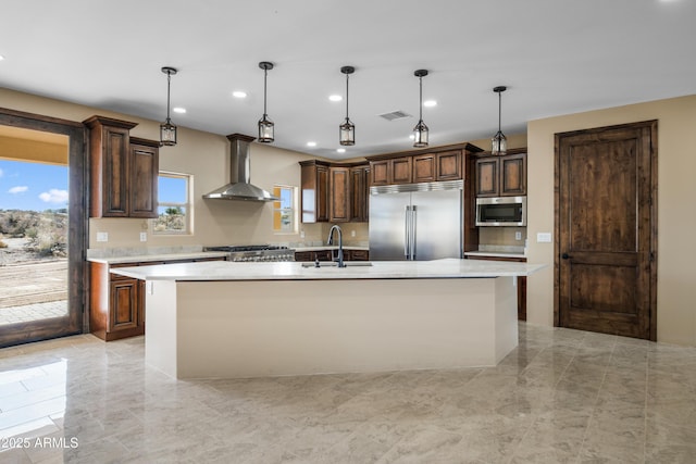 kitchen featuring pendant lighting, sink, wall chimney exhaust hood, an island with sink, and appliances with stainless steel finishes