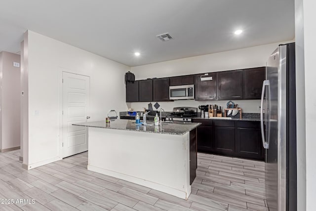 kitchen with a kitchen island with sink, dark brown cabinetry, dark stone countertops, and appliances with stainless steel finishes