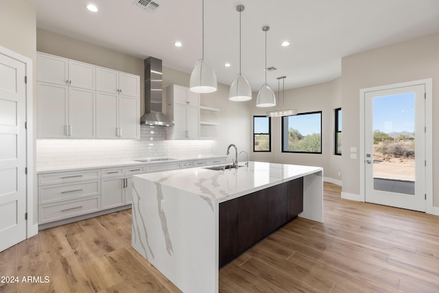 kitchen with wall chimney range hood, sink, an island with sink, black electric cooktop, and white cabinetry