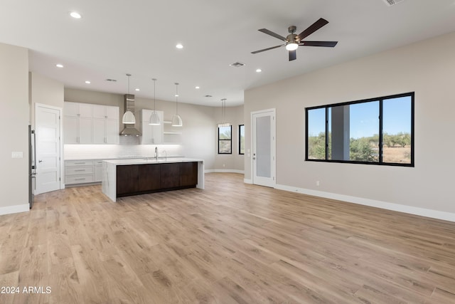 kitchen featuring white cabinets, a center island with sink, light wood-type flooring, pendant lighting, and wall chimney exhaust hood