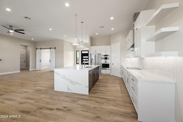 kitchen with an island with sink, hanging light fixtures, ventilation hood, a barn door, and white cabinets