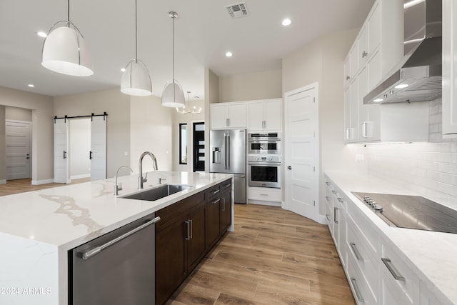 kitchen with wall chimney range hood, light wood-type flooring, a barn door, stainless steel appliances, and decorative light fixtures