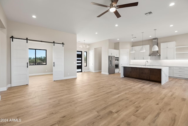 kitchen with wall chimney range hood, a kitchen island with sink, white cabinets, and a barn door