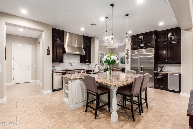 kitchen featuring wall chimney exhaust hood, an island with sink, stainless steel appliances, and backsplash