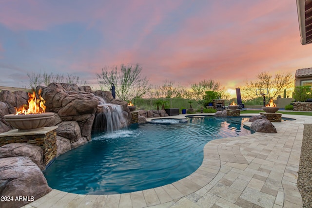 pool at dusk featuring an outdoor fire pit, an in ground hot tub, pool water feature, and a patio area