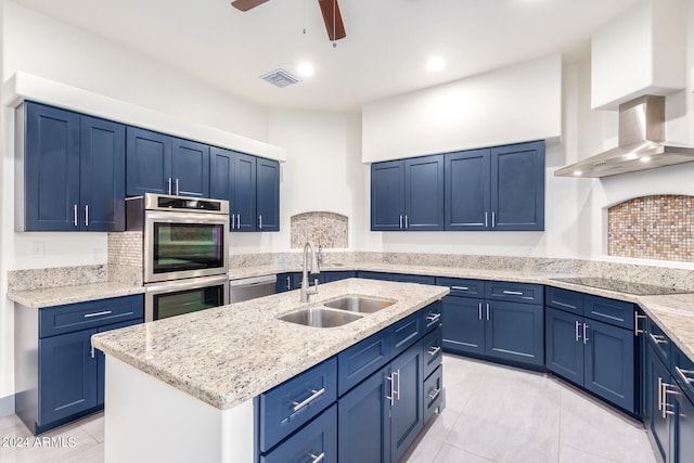 kitchen with blue cabinetry, sink, a center island with sink, appliances with stainless steel finishes, and wall chimney range hood