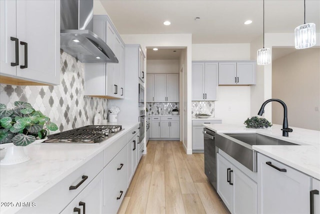 kitchen with wall chimney exhaust hood, light stone counters, appliances with stainless steel finishes, pendant lighting, and a sink