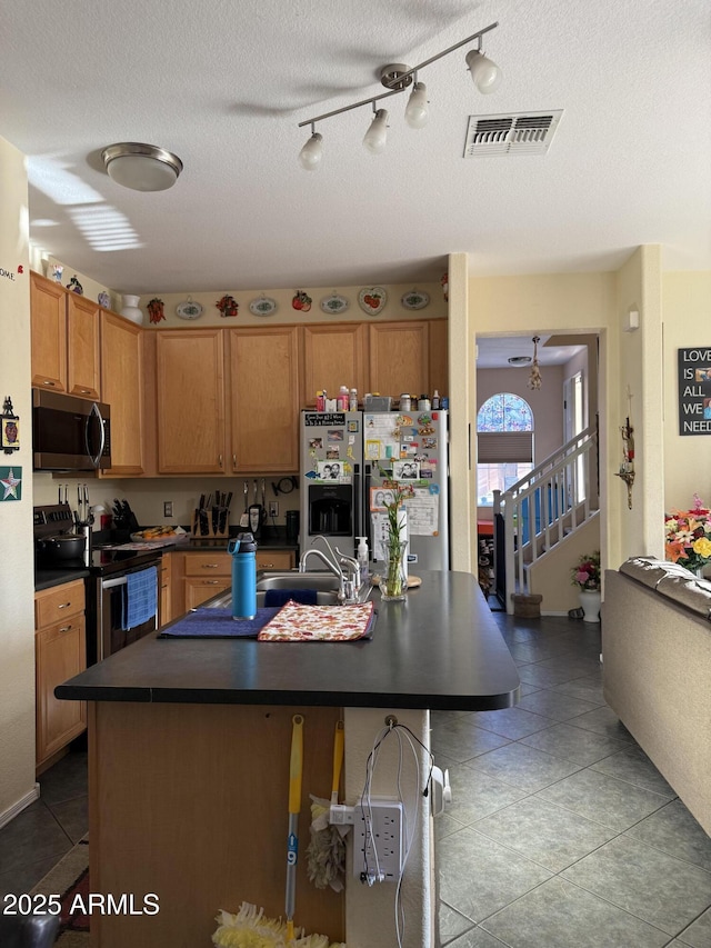 1448 E Mobile Ln, Phoenix, AZ 85040 - kitchen featuring stainless steel appliances, dark countertops, light tile patterned floors, brown cabinets, and an island with sink