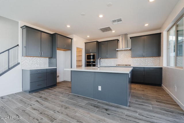 kitchen with hardwood / wood-style floors, a center island with sink, backsplash, cooktop, and wall chimney exhaust hood