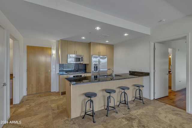 kitchen with dark stone countertops, a peninsula, stainless steel appliances, light brown cabinetry, and a sink