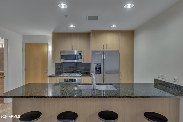 kitchen featuring kitchen peninsula, appliances with stainless steel finishes, light brown cabinetry, dark stone countertops, and sink