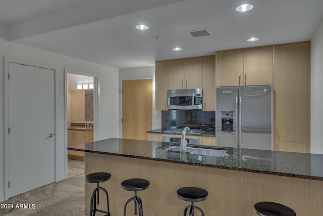 kitchen featuring stainless steel appliances, dark stone countertops, sink, a breakfast bar, and light brown cabinetry